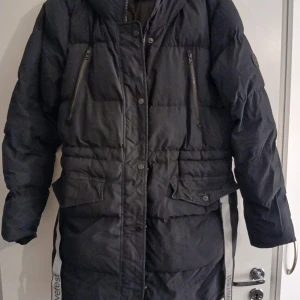 Svart lång parkas från Everest storlek 42 - Lång svart parkas från Everest med huva, dragkedja och tryckknappar framtill. Jackan har två bröstfickor med dragkedja och två stora fickor med lock. Snygga reflexdetaljer på sidorna och quiltad design för extra värme.