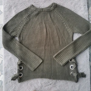 Olivgrön stickad tröja med snörning - Snygg olivgrön stickad tröja med lång ärm och coola snörningsdetaljer med metallringar i sidorna. Tröjan har en rak passform och ribbstickad struktur som ger en trendig och avslappnad look. Perfekt för lager-på-lager under hösten och vintern.