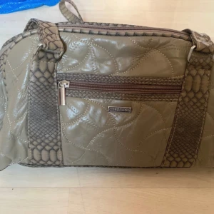 Beige axelväska från Stefano - Snygg beige axelväska från Stefano med quiltad yta och detaljer i ormskinnsmönster. Väskan har ett stort fack med innerfickor, dragkedja upptill och en yttre ficka med dragkedja. Perfekt storlek för att få plats med allt du behöver.