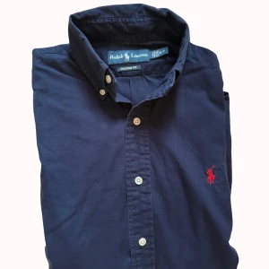 Marinblå skjorta från Ralph Lauren - Snygg marinblå skjorta från Ralph Lauren med klassisk krage och vita knappar. Custom fit-modell med lång ärm och ikonisk röd logobroderi på bröstet. Skjortan är gjord i mjuk bomull och passar perfekt till jeans eller chinos.
