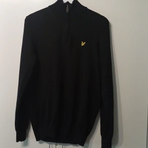 Svart stickad tröja Lyle & Scott half zip - Svart finstickad tröja från Lyle & Scott med half zip och gul logga på bröstet. Hög krage och långa ärmar. Klassisk och stilren modell som funkar året runt.