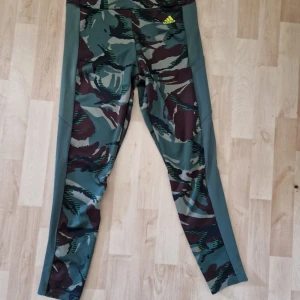 Camo leggings från Adidas Primegreen M - Snygga camo-mönstrade leggings från Adidas i storlek M. Byxorna har gröna och bruna toner med gult Adidas-tryck framtill. Tillverkade i stretchigt syntetmaterial (Primegreen, Aeroready) som andas och passar perfekt till träning.