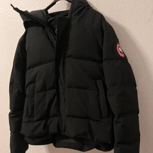 Canada goose MacMillan - Säljer en svart pufferjacka från Canada Goose med klassisk logga på ärmen. Jackan har huva, stora fickor och dragkedja framtill. Perfekt för kalla dagar och riktigt snygg streetstil. Kommer med originalpåse. Använt den max 2 gånger. Hämtas upp i Malmö , inga beställningar ! Obs; det finns ett litet sår som man enkelt kan sy , såret syns inte allt. Inga return