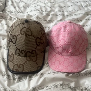 Gucci kepsar i beige och rosa - Två snygga Gucci kepsar med GG-logga över hela kepsen. Den ena är beige med bruna detaljer och svart kant, den andra är rosa med ljusrosa GG-mönster och rosa kant. Båda har justerbar rem baktill och är tillverkade i bomullsmaterial. Perfekt för dig som vill sticka ut med lyxig streetstyle.