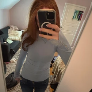 Ljusblå tight långärmad tröja - Supersnygg ljusblå långärmad tröja med tight passform och dekorativa knappar vid ärmsluten. Perfekt att styla med jeans eller kjol för en clean look. Mjuk och skön, passar både till skolan och häng med kompisar.