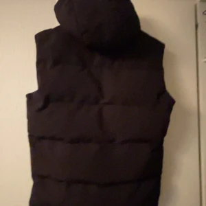 Superdry dunväst med huva - Svart dunväst från superdry med huva och tryckknappar framtill. Västen har två stora fickor och quiltad design som ger en schysst streetkänsla. 