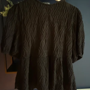 Svart blus med puffärm från Gina Tricot - Supersnygg svart blus från Gina Tricot med puffiga korta ärmar och vågig struktur i tyget. Blusen har en feminin volang nertill och en öppen rygg med knytband. Perfekt för dig som gillar trendiga detaljer och en unik look.