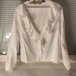 Vit blus med volang och knyt - Säljer en vit blus med djup v-ringning, volangdetaljer längs ringningen och knyt framtill. Blusen har långa ärmar och är tillverkad i ett lätt, vävt material som ger en luftig känsla. Perfekt för dig som gillar romantiska och feminina detaljer.