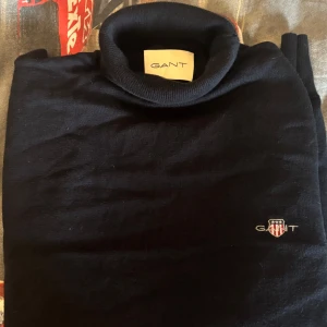 Marinblå polotröja från GANT - En marinblå polotröja från GANT som är använd en gång med broderad logga på bröstet. Tröjan är långärmad och har en ribbad polokrage. Tillverkad i mjuk bomull som känns skön mot huden. Perfekt för lager-på-lager och stilren look.