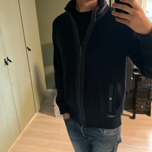 Mörkblå cardigan från Sebago - Säljer en mörkblå cardigan från Sebago med hel dragkedja framtill och ribbade muddar. Koftan har två fickor med snygga detaljer och hög krage. Perfekt för kyliga dagar och enkel att matcha med jeans eller chinos. 