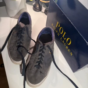 Grå mockasneakers Polo Ralph Lauren - Snygga grå sneakers från Polo Ralph Lauren i mocka med klassisk låg profil och vit sula. Skorna har svart snörning och den ikoniska broderade Polo-loggan på sidan. Perfekta för en stilren och avslappnad look.