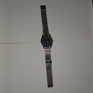 Silverfärgad klocka från Regal - Stilren klocka från Regal med svart urtavla och romerska siffror. Armbandet är i silverfärgat metallnät och har ett klassiskt spänne. Klockan har en rund form och en minimalistisk design som passar till många olika stilar.