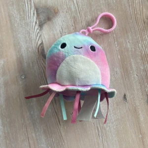 Squishmallows - Bilderna visar en söt, pastellfärgad mjukisdjur-nyckelring i form av en manet med leende ansikte och färgglada band. Produkten har en rosa plastklämma för enkel fastsättning på väska eller nycklar. Inga synliga defekter eller slitage. Perfekt som accessoar eller present.