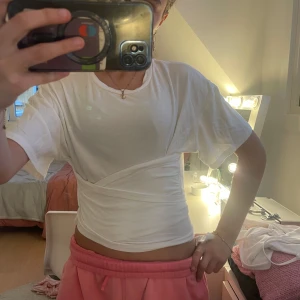 Vit omlott-topp med knyt från Cider - Säljer en vit croppad topp från Cider med kort ärm och snygg omlott-detalj som knyts i ryggen. T-shirten är i mjuk bomull och har en clean look som passar perfekt till jeans eller kjol. Enkel och stilren design med rund halsringning.