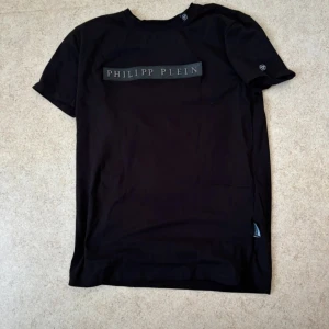 Svart Philipp Plein t-shirt Platinum Cut - Svart t-shirt från Philipp Plein med Platinum Cut, stilren logotyp framtill och diskret emblem på ärmen. Klassisk rund hals och korta ärmar. Mjuk bomullskvalitet och normal passform. Perfekt för dig som gillar exklusiva streetwear-vibbar.