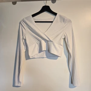 Vit omlott croppad långärmad topp - !!Finns 2st!! Säljer en vit croppad topp med omlott-design och v-ringning. Toppen är ribbad och har långa ärmar, vilket ger en snygg och modern look. Perfekt att styla med höga jeans eller kjol för en trendig outfit.