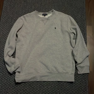 Grå sweatshirt Polo Ralph Lauren - Klassisk grå sweatshirt från Polo Ralph Lauren i storlek 170. Tröjan har rund halsringning, lång ärm och den ikoniska lilla broderade loggan på bröstet. Tillverkad i mjuk bomullsblandning med polyester för extra komfort. Vet inte varför det är storlek xl den passar 170, använd fåtal gånger nästan ny