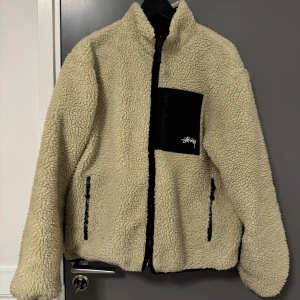 Stüssy teddyjacka med 8-ball motiv - Säljer en fet teddyjacka från Stüssy med krämfärgad utsida i teddy och svart bröstficka med logga. Baksidan har en stor svart panel med ikoniskt 8-ball motiv i vitt. Jackan är vändbar med svart utsida och broderad Stüssy-logga. Dragkedja framtill och två fickor.