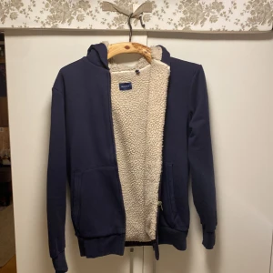Mörkblå hoodie med teddyfoder från GANT - Mysig mörkblå hoodie från GANT med dragkedja och huva. Insidan är fodrad med fluffigt, beige teddyfoder som håller dig varm. Perfekt för kyliga dagar och har två fickor framtill. Enkel och stilren design med klassisk passform. Använd fåtal gånger och är i mycket bra skick 