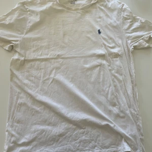 Vit t-shirt från Ralph Lauren - Vit t-shirt från Ralph Lauren med klassisk passform och korta ärmar. T-shirten har en liten mörkblå broderad logga på bröstet och är tillverkad i mjuk bomull. Perfekt basic-plagg med stilren look.