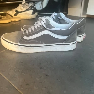 Gråa Vans Old Skool sneakers - Säljer ett par klassiska Vans Old Skool sneakers i grått med vit sula och vit sidostrip. Skorna har snörning och är tillverkade i en mix av mocka och canvas. Perfekta för dig som gillar en clean och tidlös streetstil.