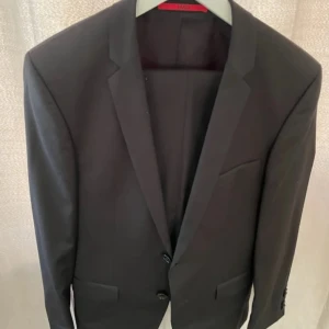Svart kavaj från Hugo Boss, strl 44 - Säljer en klassisk svart kavaj från Hugo Boss i storlek 44. Kavajen har två knappar framtill, fickor med lock och smal krage. Levereras med original klädpåse från Boss. Perfekt för en stilren look. Använd i några timmar vid ett tillfälle.