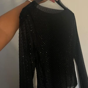 Svart mesh topp med glitterprickar - Säljer en svart mesh topp med långa ärmar och glittriga små prickar över hela tyget. Toppen är genomskinlig och har en tight passform som sitter snyggt på kroppen. 