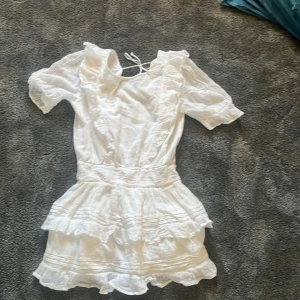 Vit kortklänning med volanger Gina Tricot - Supersöt vit kortklänning från Gina Tricot med volanger och smock i midjan. Klänningen har korta puffärmar, v-ringning med knyt i nacken och flera lager volanger på kjolen. Perfekt för sommarens alla tillfällen!