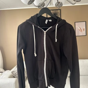 Svart hoodie med dragkedja från Icon - Svart hoodie från Icon med vit dragkedja och vita snören i huvan. Klassisk modell med känguruficka framtill och ribbade muddar vid ärmslut och nederkant. Perfekt för chill dagar eller när du vill ha en enkel streetstyle-look.