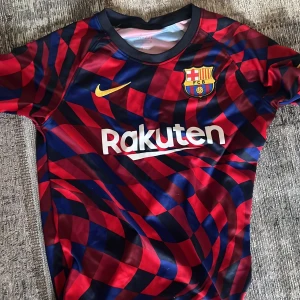 FC Barcelona Nike fotbollströja röd/blå  - Säljer en FC Barcelona fotbollströja från Nike med klubbmärke på bröstet och Rakuten/Beko-tryck. Tröjan har ett unikt röd- och blåmönster, korta ärmar och är tillverkad i lätt, ventilerande polyester. Perfekt för dig som älskar Barca och vill sticka ut på planen.