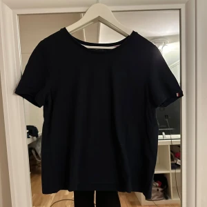 Mörkblå t-shirt från Tommy Hilfiger - Mörkblå t-shirt från Tommy Hilfiger med broderad TH-logga på bröstet och en liten flaggdetalj på ärmen. Klassisk rund halsringning och rak passform. Tillverkad i mjuk bomull som känns skön mot huden.