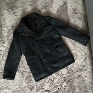 Svart jacka ZARA - Stilren svart skepparkavaj med dubbelknäppning och bred krage i fuskpäls. Jackan har flera fickor framtill och klassisk rak passform. Perfekt för dig som vill ha en tidlös och snygg look under kyliga dagar.