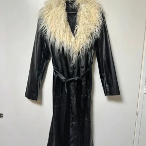Svart lång skinnrock med päls - Lång svart skinnrock  med stor fluffig vit päls runt kragen. Jackan har bälte i midjan och långa ärmar, vilket ger en riktigt statement look. Perfekt för dig som vill sticka ut med en cool och unik stil. Aldrig använd Längd 117.5cm.
