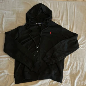 Svart hoodie från Polo Ralph Lauren - Svart hoodie från Polo Ralph Lauren med dragkedja och huva. Klassisk röd broderad logga på bröstet och snörning i huvan. Tillverkad i mjuk bomull, perfekt för chill dagar. Custom fit-modell med ribbade muddar och fickor framtill. Bara använd enstaka gång. Undviker skambud. Finns att hämta utanför Jönköping om så önskas. 