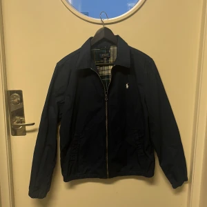 Marinblå vindjacka från Polo Ralph Lauren - Snygg marinblå vindjacka från Polo Ralph Lauren med klassisk krage, dragkedja och broderad logga på bröstet. Jackan har rutigt innerfoder och elastiska muddar vid ärmslut och midja. Perfekt för dig som gillar stilren och tidlös design. Storlek XL för junior (runt 150cm) ny pris 1900kr, knappt använd. Pris kan diskuteras.