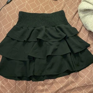 Svart volangkjol från Vero Moda - Supersöt svart kortkjol från Vero Moda med bred smock i midjan och flera lager volanger. Kjolen är perfekt för dig som gillar en feminin och trendig look. Lätt och luftig känsla, passar snyggt till både sneakers och boots.