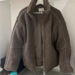 Brun teddyjacka med dragkedja - Mysig brun teddyjacka med hög krage och grov dragkedja framtill. Jackan har en loose passform och är tillverkad i fluffigt teddy-material som håller dig varm. Perfekt för kyliga dagar och har en enkel, stilren look.