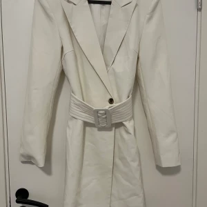 Vit blazerklänning från Vila - Snygg vit blazerklänning från Vila med markerade axlar och lång ärm. Klänningen har en bred, quiltad bälte i midjan och djup v-ringning. Stilren design med knappstängning framtill, perfekt för dig som gillar minimalistisk och chic look.