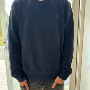 Svart sweatshirt från Polo Ralph Lauren - Svart sweatshirt från Polo Ralph Lauren med klassisk röd broderad logga på bröstet. Tröjan har rund hals, långa ärmar och mjukt bomullsmaterial som är skönt mot huden. Perfekt för chill dagar och enkel att matcha med jeans eller joggers.