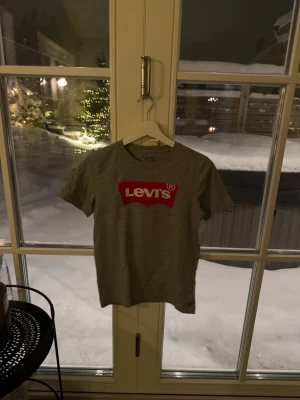 Grå Levi's t-shirt med röd logga - Säljer en grå t-shirt från Levi's med den klassiska röda loggan tryckt på bröstet. T-shirten har rund hals och korta ärmar. Tillverkad i mjuk bomull som känns skön mot huden. Perfekt för en avslappnad och stilren look.