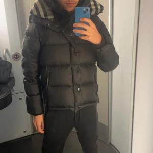 Svart dunväst/jacka med rutigt foder - Säljer en svart dunväst med knappar och dragkedja. Västen och jackan  har ett rutigt foder i huvan med grå, vit och röd färg. Perfekt för lager-på-lager och har en klassisk, puffad look. Jackan kommer ifrån märket Burberry london England. Man kan ta av armarna så att du kan ha på dig den på sommar kvällerna. 