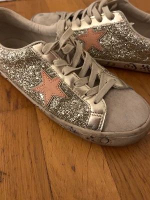 Glittriga sneakers med stjärndetalj - Säljer ett par sneakers från Next med silverglittriga sidor och en rosa stjärna på varje sida. Det är unika o fina o i bra skick använd runt 5 gånger men tyvärr för små 