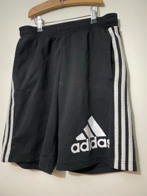 Svarta Adidas shorts med vita ränder - Säljer ett par klassiska svarta Adidas shorts med vita ränder längs sidorna och stort Adidas-tryck på ena benet. De har elastisk midja och är gjorda i mjuk bomull, perfekta för träning eller chill. Snygg och sportig look som alltid funkar.