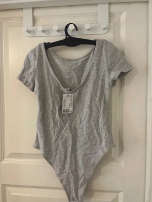 Grå basic body från H&M - Säljer en enkel och stilren grå body från H&M Basic. Bodyn har korta ärmar, rundad urringning och är tillverkad i mjuk bomullsmix. Perfekt att styla med jeans eller kjol för en clean look.