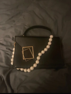 Svart handväska med pärlhandtag - Säljer en svart handväska i skinnimitation med ett unikt handtag av stora vita pärlor och en cool guld-detalj framtill. Väskan har en stilren, rektangulär form och ett kort bärhandtag upptill. Perfekt för dig som vill sticka ut med accessoarer.