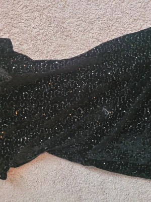 Svart paljettklänning i sammet - Säljer en svart sammetsklänning täckt med glittriga paljetter. Klänningen har en hög hals och är figurnär. Materialet är mjukt och skimrande, vilket ger en lyxig känsla.