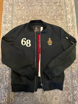 Svart bomberjacka från Morris - Svart bomberjacka från Morris med broderad logga och siffran 68 på bröstet. Jackan har dragkedja framtill, ribbade muddar och en Union Jack-flagga som foder. Snygg detalj med ficka på ärmen och klassisk bomberstil.