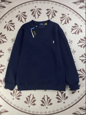Mörkblå sweatshirt från Polo Ralph Lauren - Snygg mörkblå sweatshirt från Polo Ralph Lauren med klassisk rund hals och vit broderad logga på bröstet. Tröjan har ribbade muddar vid ärmslut och nederkant, samt en stilren och enkel design som passar till många olika outfits.