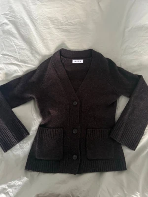 Mörkbrun kofta från Wera - Snygg mörkbrun kofta från Wera med ribbade muddar och stora framfickor. Koftan har v-ringad hals, tre knappar framtill och är stickad i mjukt material. Perfekt för lager-på-lager under hösten och vintern. 100% ULL