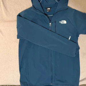  The North Face Blå vindjacka  - Blå vindjacka från The North Face med huva och hel dragkedja framtill. Jackan har ett stilrent rutmönstrat tyg på axlar och bröst. Regn tålig. Ny Pris 1400. Pris går att diskutera vid snabbt köp